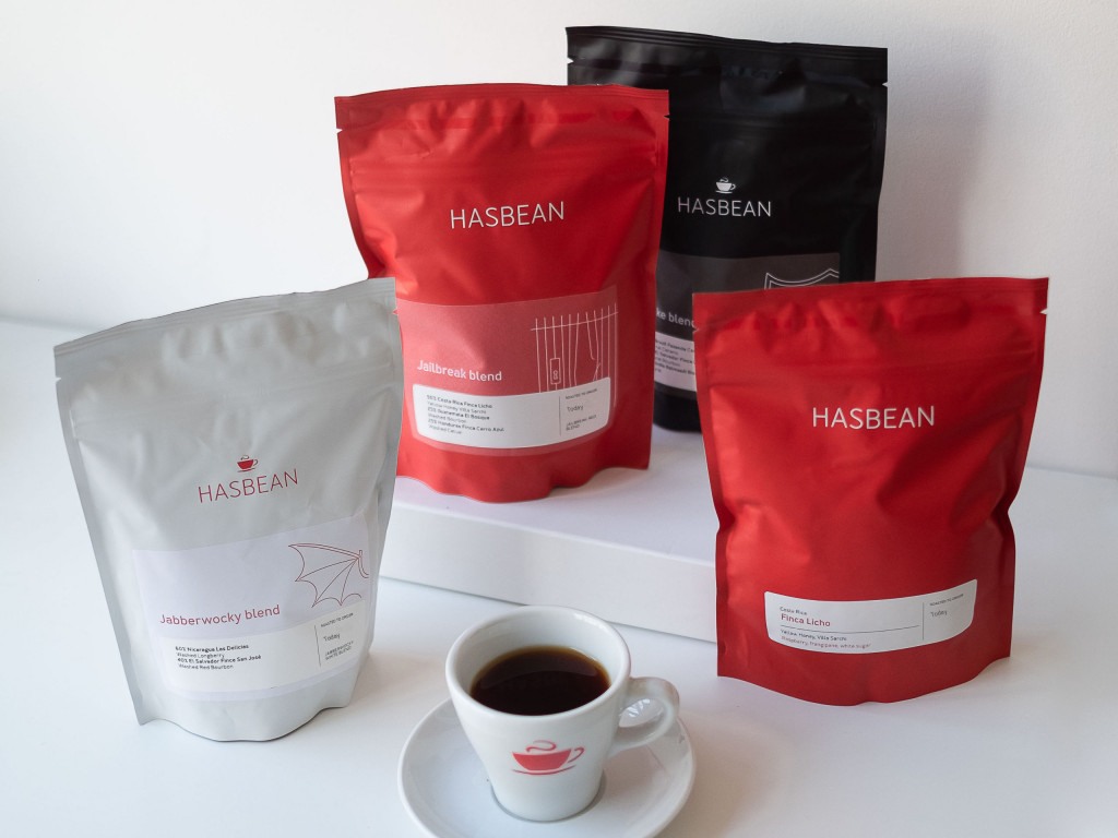 dva červené, jeden bílý a jeden černý balíček kávy od pražírny Hasbean a šálek filtrované kávy